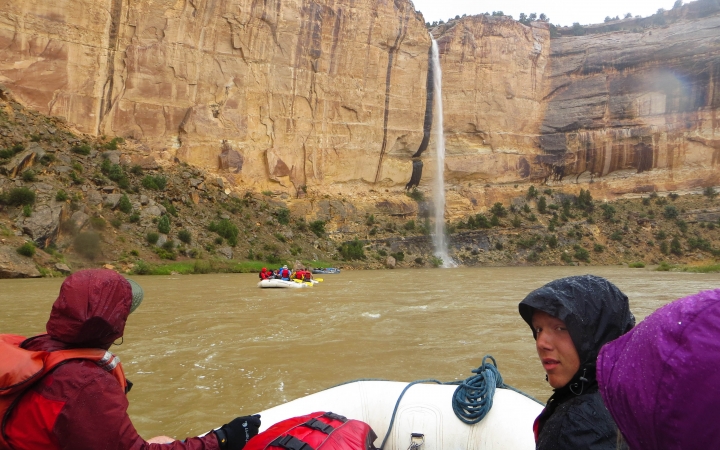 From the back of a raft, students are seen sitting in front of the raft. Beyond them, another raft floats on the water towards a waterfall.